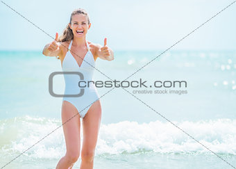 Happy young woman in swimsuit on sea shore showing thumbs up