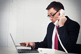 businessman speaking on phone, sitting at desk, looking at lapto