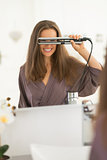 Happy young woman looking through hair straightener in bathroom