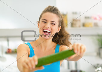 Happy young woman in kitchen showing cucumber