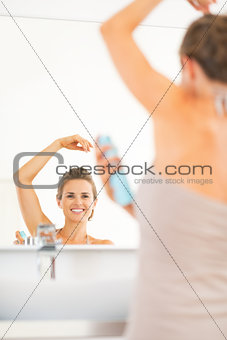 Happy young woman applying deodorant on underarm in bathroom
