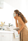Young woman wearing facial cosmetic mask in bathroom looking in 