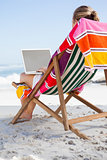 Woman sitting on beach in deck chair using laptop