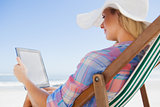 Woman in sunhat sitting on beach in deck chair using tablet pc