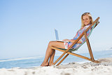 Blonde sitting on beach using her laptop smiling at camera