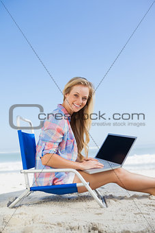 Pretty blonde sitting on beach using her laptop smiling at camera