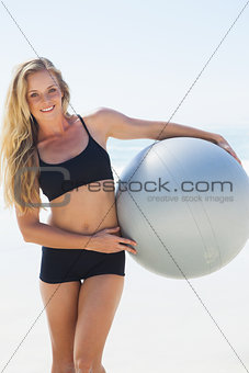 Fit blonde holding exercise ball at the beach smiling at camera