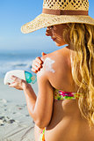 Gorgeous woman sitting on the beach in sunhat applying suncream