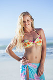 Pretty blonde in bikini and sarong smiling at camera on the beach