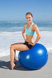 Fit woman sitting on exercise ball at the beach smiling at camera
