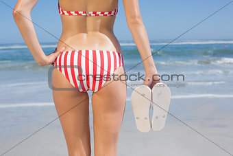 Mid section of fit woman in bikini on the beach holding flip flops
