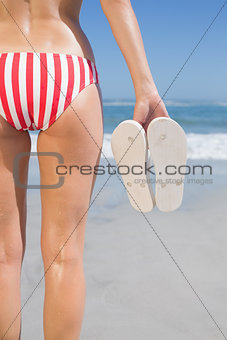 Mid section of fit woman in bikini on the beach holding flip flops