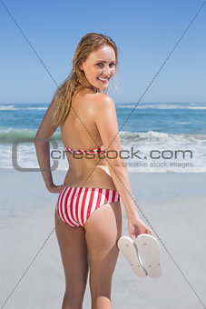 Smiling fit woman in bikini on the beach holding flip flops