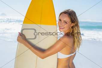 Smiling blonde surfer in white bikini holding her board on the beach