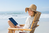 Smiling blonde sitting on wooden deck chair by the sea using tablet
