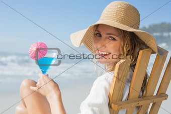 Smiling blonde relaxing in deck chair by the sea holding cocktail