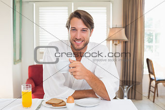 Handsome man having breakfast in his bathrobe smiling at camera
