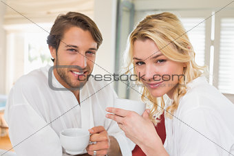 Cute couple in bathrobes having coffee together smiling at camera