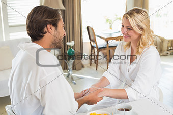 Cute couple in bathrobes having breakfast together holding hands