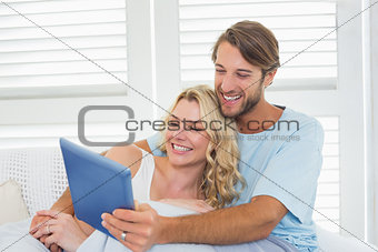 Smiling casual couple sitting on couch under blanket using tablet pc
