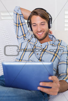 Casual smiling man lying on couch listening to music on tablet pc