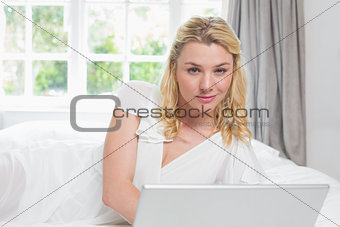 Pretty blonde lying on bed using laptop smiling at camera