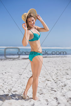 Slender woman in bikini on beach wearing sunhat smiling at camera