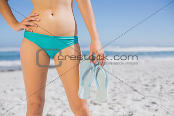 Mid section of fit woman in bikini on beach holding flip flops