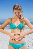 Fit smiling woman in bikini on the beach making heart shape on stomach