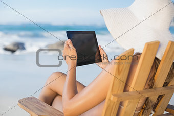 Woman in straw hat relaxing in deck chair on the beach using tablet pc