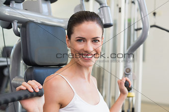 Fit brunette using weights machine for arms smiling at camera