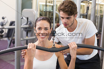 Fit brunette using weights machine for arms with trainer helping smiling at camera