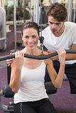 Fit brunette using weights machine for arms with trainer helping smiling at camera