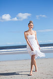 Blonde in white dress walking on the beach smiling at camera