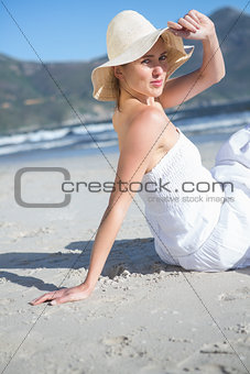 Content blonde in white dress sitting on the beach looking to camera