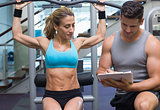 Female bodybuilder using weight machine for arms with trainer taking notes