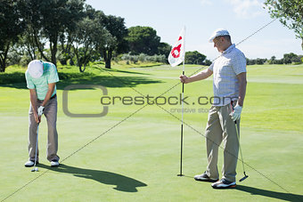 Golfer holding eighteenth hole flag for friend putting ball