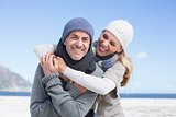 Attractive couple hugging and smiling at camera on the beach in warm clothing