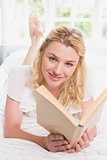 Pretty blonde lying on bed smiling at camera with book