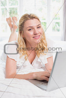 Pretty blonde lying on bed using laptop smiling at camera