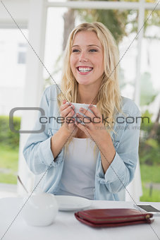 Pretty blonde sitting at table having coffee smiling at camera