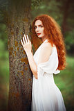Beautiful redhead woman wearing white dress stands near tree