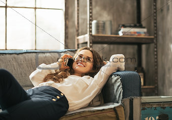 Happy young woman laying on sofa and talking cell phone in loft 