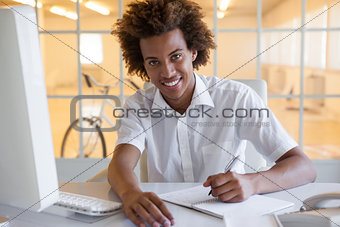 Casual young businessman writing at his desk smiling at camera