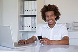 Casual smiling businessman using his smartphone and laptop at his desk
