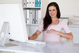 Casual pregnant businesswoman touching her bump at her desk smiling at camera