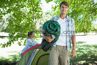 Handsome young man smiling at camera while girlfriend pitches tent