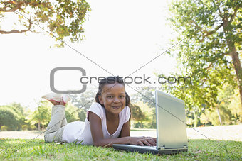 Little girl lying on grass using laptop smiling at camera