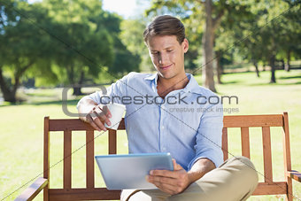 Smiling man sitting on park bench using tablet drinking coffee