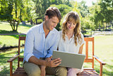 Cute couple sitting on park bench together looking at laptop